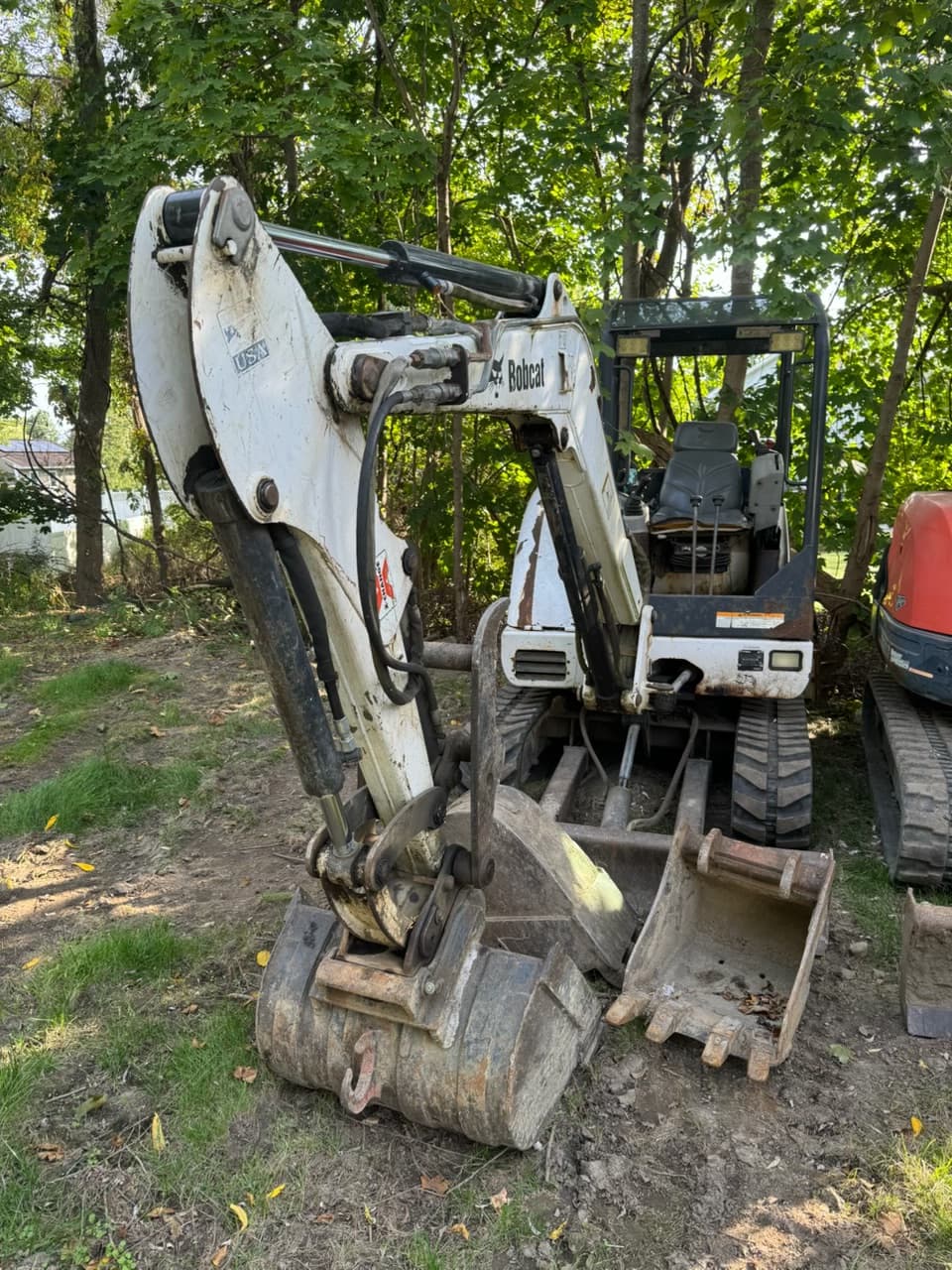 White Bobcat mini excavator with its bucket lowered, parked on dirt near a wooded area.